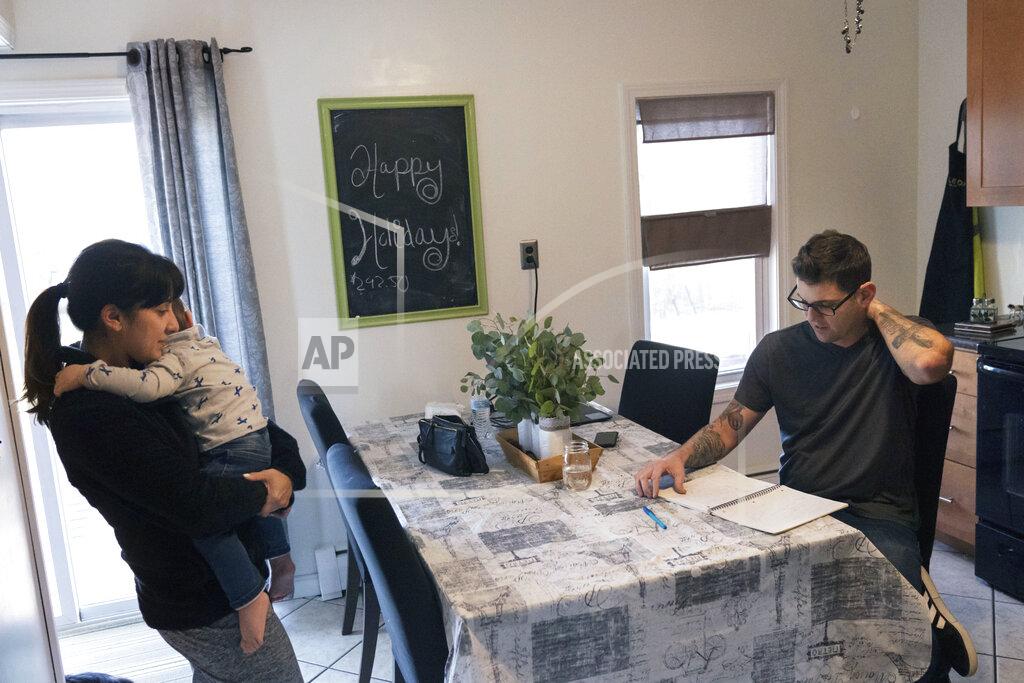 Comedian Brad Pierce, right, looks over the notebook us uses to write jokes as he sits in the kitchen next to his wife, Carmen, and their nephew, Maddox, 1, in West Warwick, R.I., Friday, Jan. 8, 2021. Pierce was finally doing well with his comedy when the pandemic hit. Now he wonders if he can possibly build up his career again. He has a friend who drives for Amazon and fears having to get a job like that while talking about the good old days when he was an entertainer. (AP Photo/David Goldman)