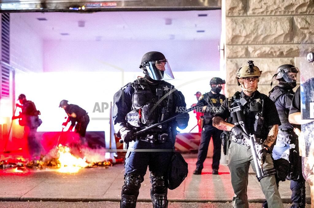 Noah Berger FILE - Police officers stand watch as fellow officers extinguish a fire lit by protesters behind the Mark O. Hatfield Federal Courthouse, Aug. 2, 2020, in Portland, Ore. Following an agreement between Democratic Gov. Kate Brown and the Trump administration to reduce federal officers in the city, nightly protests remained largely peaceful without major confrontations between protesters and officers. (AP Photo/Noah Berger, File)