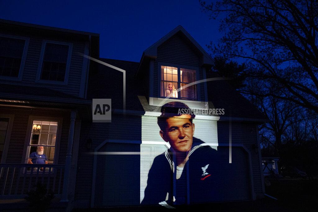 David Goldman An image of veteran Stephen Kulig is projected onto the home of his daughter, Elizabeth DeForest, as she looks out the window of a spare bedroom as her husband, Kevin, sits downstairs in Chicopee, Mass., Sunday, May 3, 2020. Kulig, a U.S. Navy veteran and resident of the Soldier's Home in Holyoke, Mass., died from the COVID-19 virus at the age of 92. After saying goodbye to her father for the last time in person, Elizabeth slept in the spare bedroom upstairs for two weeks as a precaution against possibly infecting her husband. Seeking to capture moments of private mourning at a time of global isolation, the photographer used a projector to cast large images of veterans on to the homes as their loved ones are struggling to honor them during a lockdown that has sidelined many funeral traditions. (AP Photo/David Goldman)