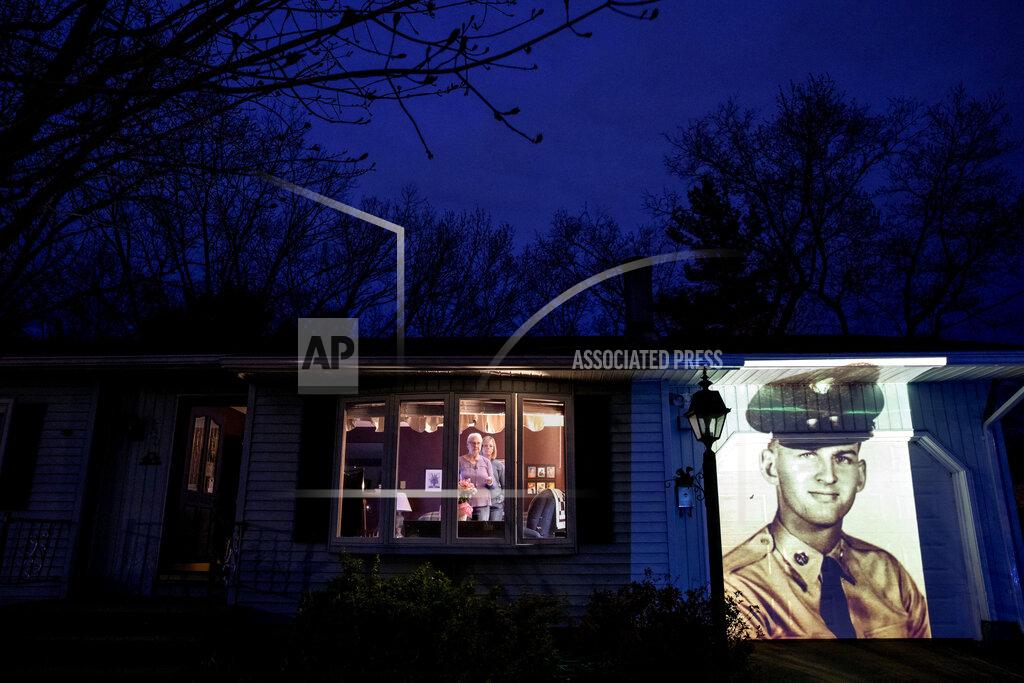 David Goldman An image of veteran Francis Foley is projected onto the home of his wife, Dale Foley, left, as she looks out a window with their daughter, Keri Rutherford, in Chicopee, Mass., Wednesday, April 29, 2020. Foley, a U.S. Army veteran and resident of the Soldier's Home in Holyoke, Mass., died from the COVID-19 virus at the age of 84. Seeking to capture moments of private mourning at a time of global isolation, the photographer used a projector to cast large images of veterans on to the homes as their loved ones are struggling to honor them during a lockdown that has sidelined many funeral traditions. (AP Photo/David Goldman)