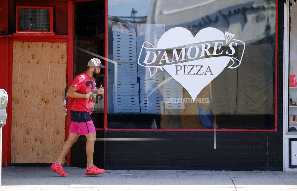 In this Monday, June 1, 2020 photo, an early morning jogger runs past a bordered D'Amores Pizza in the upscale Beverly Grove neighborhood of Los Angeles. After Bobby Alt's pizza shop was crippled for two months by the coronavirus pandemic, he reopened Saturday excited to finally dish out slices to customers in person. By the time he closed for the night, he feared the worst for his little business. A largely peaceful protest earlier in the day, which had delivered a steady flow of hungry customers, had given way to unruly groups of vandals tagging and breaking into businesses including his pizza shop. (AP Photo/Richard Vogel)