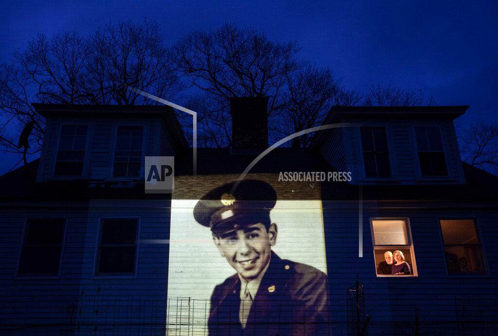 David Goldman An image of veteran Emilio DiPalma, is projected onto the home of his daughter, Emily Aho, left, as she looks out a window with her husband, George, in Jaffrey, N.H., Thursday, April 30, 2020. DiPalma, a U.S. Army WWII veteran and resident of the Soldier's Home in Holyoke, Mass., died from the COVID-19 virus at the age of 93. Seeking to capture moments of private mourning at a time of global isolation, the photographer used a projector to cast large images of veterans on to the homes as their loved ones are struggling to honor them during a lockdown that has sidelined many funeral traditions. (AP Photo/David Goldman)