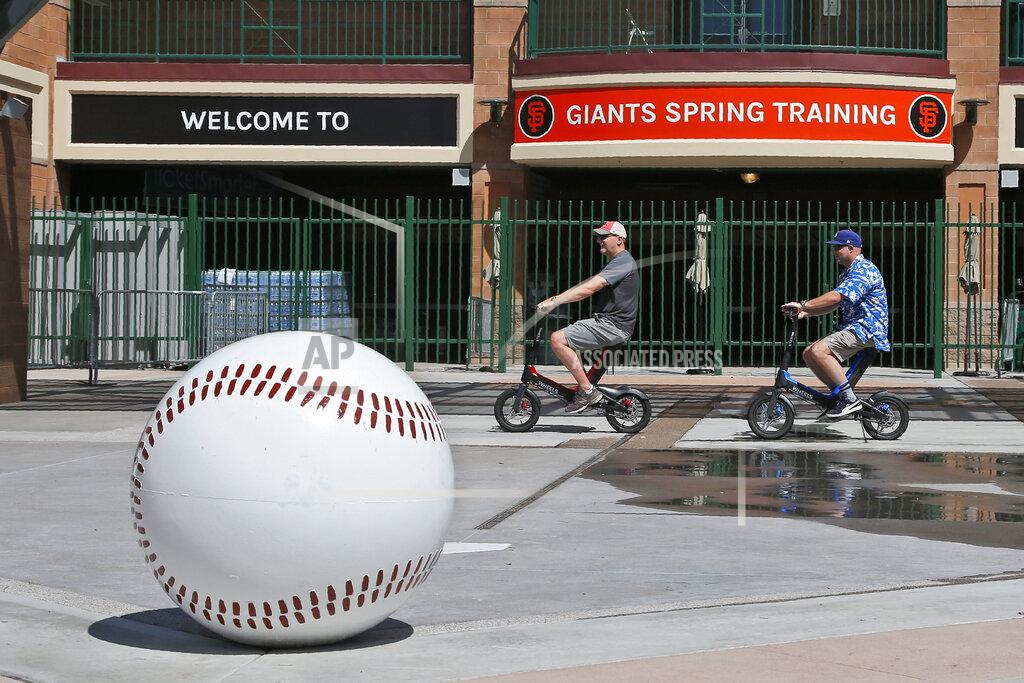 Sue Ogrocki FILE - Matt Kirmer, left, and Joe Lieser, right, both of Torrance, California, ride scooters past Scottsdale Stadium, the spring training home of the San Francisco Giants, in Scottsdale, Ariz., in this Saturday, March 14, 2020, file photo. Big league managers say that Major League Baseball has instructed them to prepare for spring training to start on time in mid-February despite uncertainty around the coronavirus. Tampa Bay Rays manager Kevin Cash said Wednesday, Dec. 16, 2020, that officials from the commissioner's office had a meeting with managers Tuesday an expressed optimism about opening spring camps as scheduled. (AP Photo/Sue Ogrocki, File)