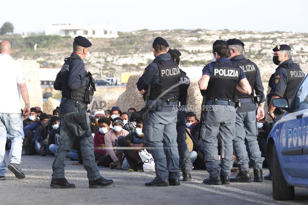 Salvatore Cavalli Migrants wearing face masks to curb the spread of COVID-19 sit at a pier as Italian police officers stand by, on the Sicilian island of Lampedusa, southern Italy, Monday, May 10, 2021. Several hundred more migrants reached the tiny Italian island before dawn on Monday, swelling to past 2,100 the number of arrivals in around 24 hours and fueling calls from across the political spectrum for the Italian government to strengthen its migration policies. (AP Photo/Salvatore Cavalli)