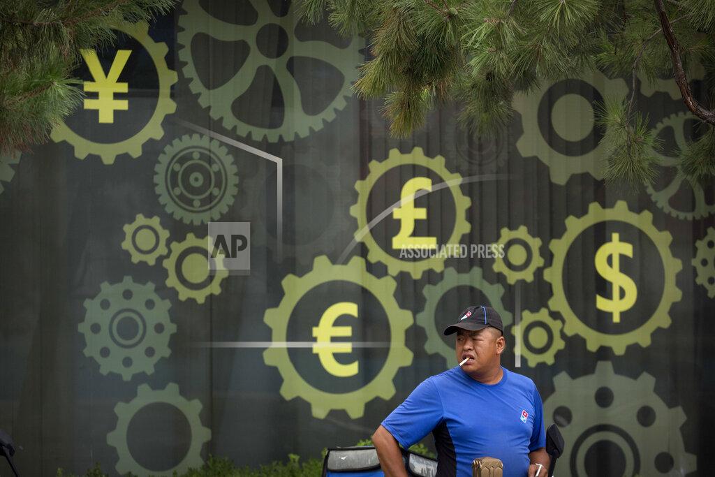 Mark Schiefelbein FILE - In this July 20, 2018, file photo, a deliveryman stands near a mural displaying Chinese yuan and other world currency symbols on the outside of a bank in Beijing. The Tokyo Olympics are already the most expensive Summer Games on record with costs set to go higher, a wide-ranging study from Britain's University of Oxford indicates. This is even before the costs of the one-year delay from the COVID-19 pandemic are known with the cost overrun already at over 200%, lead author Bent Flyvbjerg explained in an interview with the Associated Press. (AP Photo/Mark Schiefelbein, File)