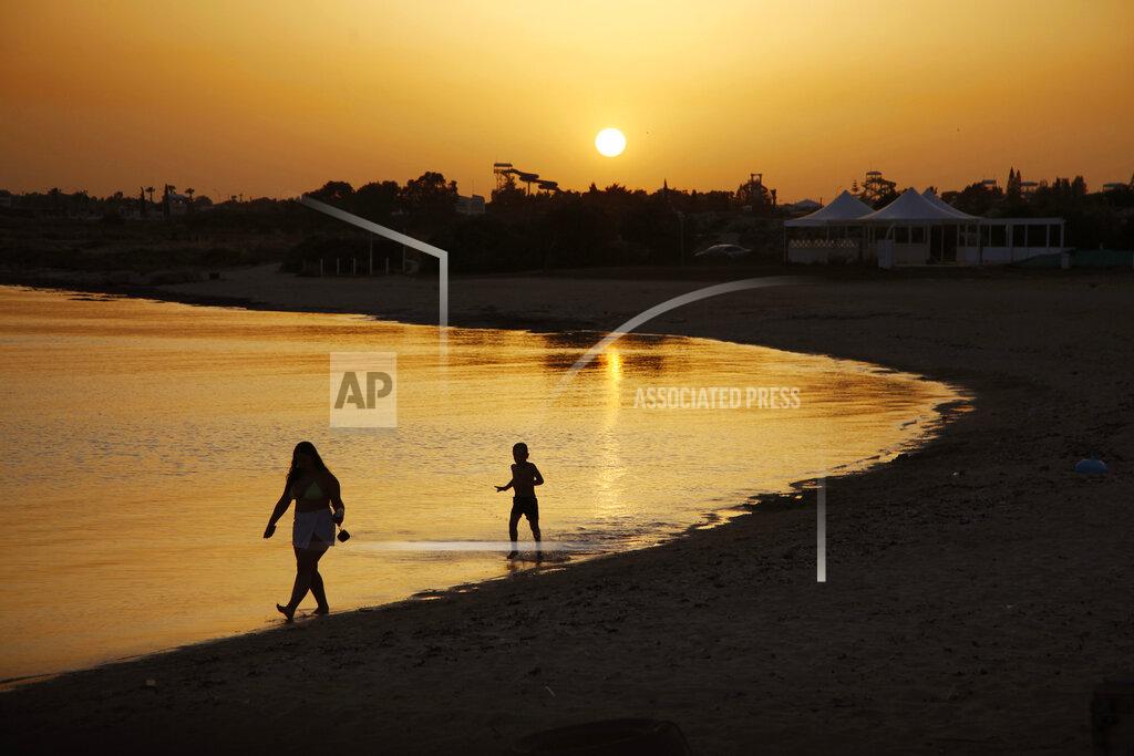 Petros Karadjias FILE - In this file photo dated Sunday, May 17, 2020, beachgoers walk in the sea water during sunset at an empty stretch of Dome beach at Makrinissos in Cyprus' seaside resort of Ayia Napa, a favorite among tourists. Cyprus' government Wednesday May 27, 2020, is pledging to cover all costs for anyone testing positive for the coronavirus while on vacation in the east Mediterranean island nation, covering the costs of lodging, food, drink and medication for COVID-19 patients and their families. (AP Photo/Petros Karadjias, FILE)