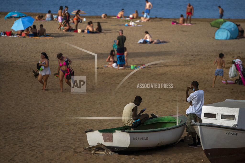 Emilio Morenatti Two migrants sit on a fishing boat as people enjoy the beach in Gran Canaria island, Spain, on Friday, Aug. 21, 2020. Though migrant arrivals to mainland Spain via the Mediterranean Sea have decreased by 50% compared to last year, landings in the Canary Islands have increased by 550% and haven’t been this high in over a decade, raising alarms at the highest levels of the Spanish government. (AP Photo/Emilio Morenatti)