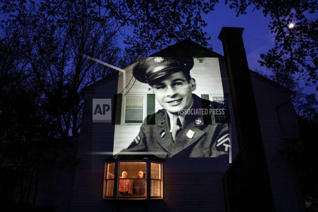 David Goldman An image of veteran James Sullivan is projected onto the home of his son, Tom Sullivan, left, as he looks out a window with his brother, Joseph Sullivan, in South Hadley, Mass., Monday, May 4, 2020. Sullivan, a U.S. Army WWII veteran and resident of the Soldier's Home in Holyoke, Mass., died from the COVID-19 virus four days shy of his 100th birthday. Seeking to capture moments of private mourning at a time of global isolation, the photographer used a projector to cast large images of veterans on to the homes as their loved ones are struggling to honor them during a lockdown that has sidelined many funeral traditions. (AP Photo/David Goldman)
