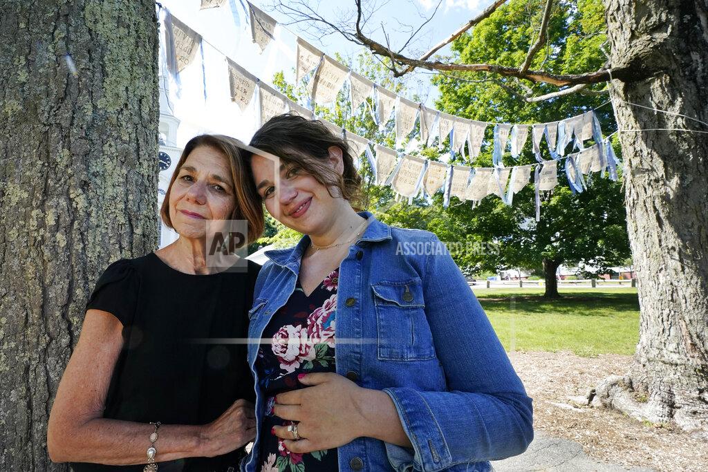 Elise Amendola Widow Marcy Jacobs, left, stands with her daughter, Jaclyn Winer, under flags bearing names of people, including her husband, Keith Jacobs, who have died from COVID-19, outside the First Congressional Church, Thursday, June 17, 2021, in Holliston, Mass. The flags are part of the COVID Art and Remembrance project spearheaded by Jaclyn. “Don’t expect us to move on without giving us a place to grieve,” Marcy Jacobs said, recalling her husband as kind, uncomplaining and simple. “Is it a stone for everyone? I don’t know." (AP Photo/Elise Amendola)