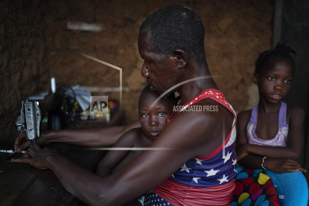 Leo Correa Osman works on his sewing machine as a sister of Marie stands with him in Komao village, on the outskirts of Koidu, district of Kono, Sierra Leone, Sunday, Nov. 22, 2020. "In the past months things were hard for me, even to have food to eat", says Marie's stepfather. (AP Photo/Leo Correa)