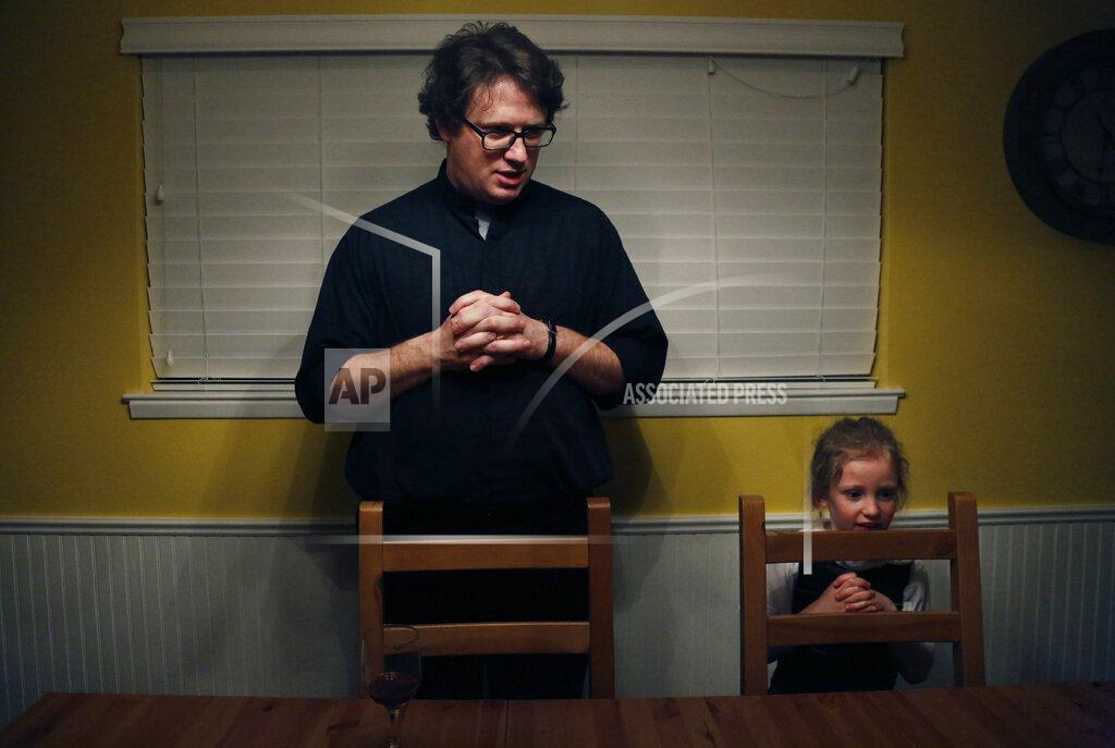 In this Feb. 7, 2020, photo, converted Catholic Priest Joshua Whitfield, left, his daughter Zoe-Catherine, 5, and the rest of his family stand behind their chairs to say grace before dinner in north Dallas. (AP Photo/Jessie Wardarski)