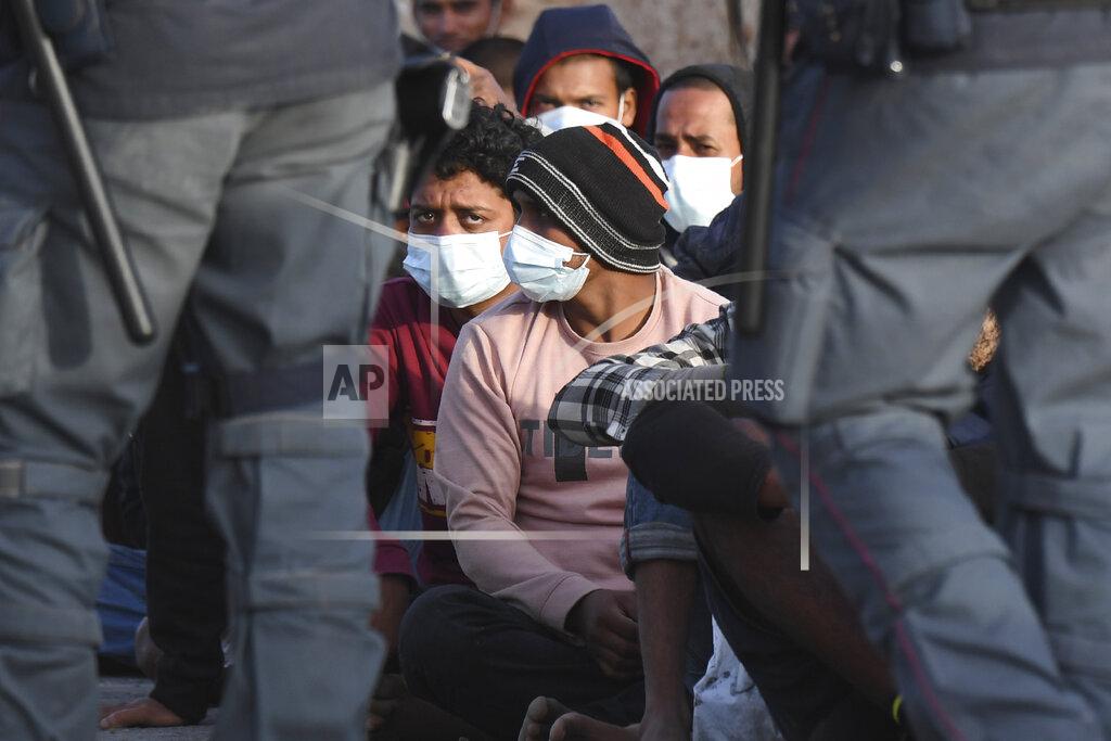 Salvatore Cavalli Migrants wearing face masks to curb the spread of COVID-19 sit at a pier as Italian police officers stand by, on the Sicilian island of Lampedusa, southern Italy, Monday, May 10, 2021. Several hundred more migrants reached the tiny Italian island before dawn on Monday, swelling to past 2,100 the number of arrivals in around 24 hours and fueling calls from across the political spectrum for the Italian government to strengthen its migration policies. (AP Photo/Salvatore Cavalli)
