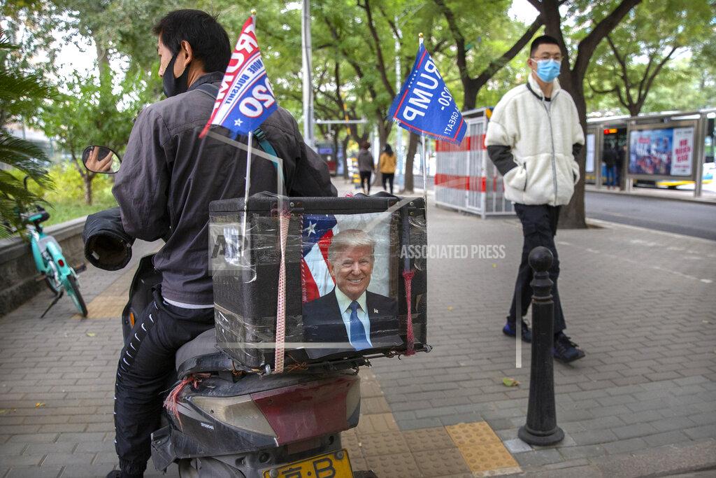 Mark Schiefelbein FILE - In this Oct. 14, 2020 file photo, a delivery courier rides a motorbike decorated with a portrait of U.S. President Donald Trump and "Trump 2020" flags in Beijing. Tuesday's U.S. election, what's at stake if Donald Trump's presidency ends — or if it continues? Nation by nation, how is Election Day in the United States being watched, considered, assessed? It's all about trade for China — but trade is about hitting economic growth targets at home and being a technology leader abroad. The stormy commercial relationship between the world's two biggest economies since President Donald Trump took office is front and center in China's view of the U.S. election. (AP Photo/Mark Schiefelbein, File)