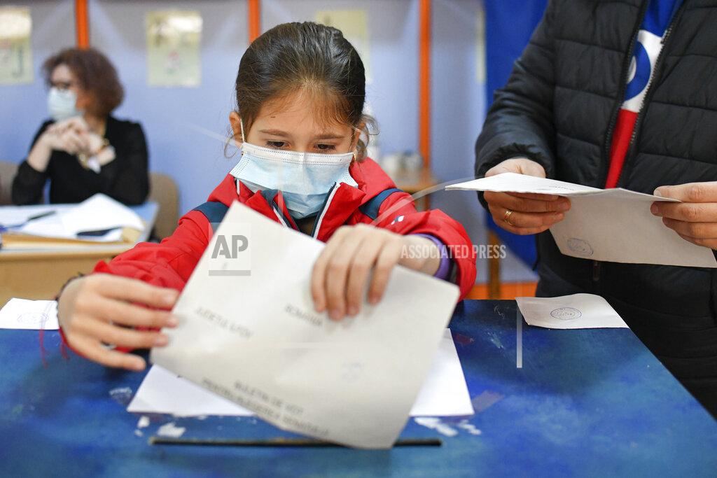 Andreea Alexandru A child wearing a mask for protection against the COVID-19 infection places an adult's vote into the ballot box in the village of Branesti, Romania, Sunday, Dec. 6, 2020. Voting started in Romania's legislative election expected to restore some measure of stability after five years of political and social turbulence with more than 18 million Romanians registered to vote for a new legislative body. (AP Photo/Andreea Alexandru)