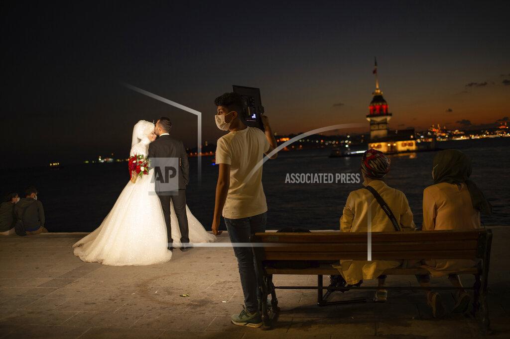 Yasin Akgul A wedding photographer's assistant, wearing a mask for protection against the spread of coronavirus, holds lighting equipment, as a couple poses for photographs backdropped by Istanbul's iconic Maiden's Tower (Kiz Kulesi) at the Bosphorus Strait separating the European and Asian sides of Istanbul, Friday, Sept. 11, 2020. Turkey is getting tough on people who flout self-isolation rules despite testing positive for the coronavirus. An Interior Ministry circular sent to the country's 81 provinces on Friday said people caught leaving their homes despite isolation orders will be quarantined and supervised at state-owned dormitories or hostels. (AP Photo/Yasin Akgul)