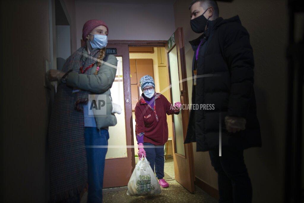 Alexander Zemlianichenko Russian pensioner Galina Prokopieva, 72, wearing a face mask to protect against coronavirus, picks up a bag with products brought by the volunteers, also wearing face masks and observe social distancing guidelines wait for an elevator in Moscow, Russia, Sunday, Jan. 3, 2021. In the framework of Charitable action "New Year's Volunteers" of the "We Are Together" movement volunteers distribute food, medicine and other necessary things to elderly people. (AP Photo/Alexander Zemlianichenko)