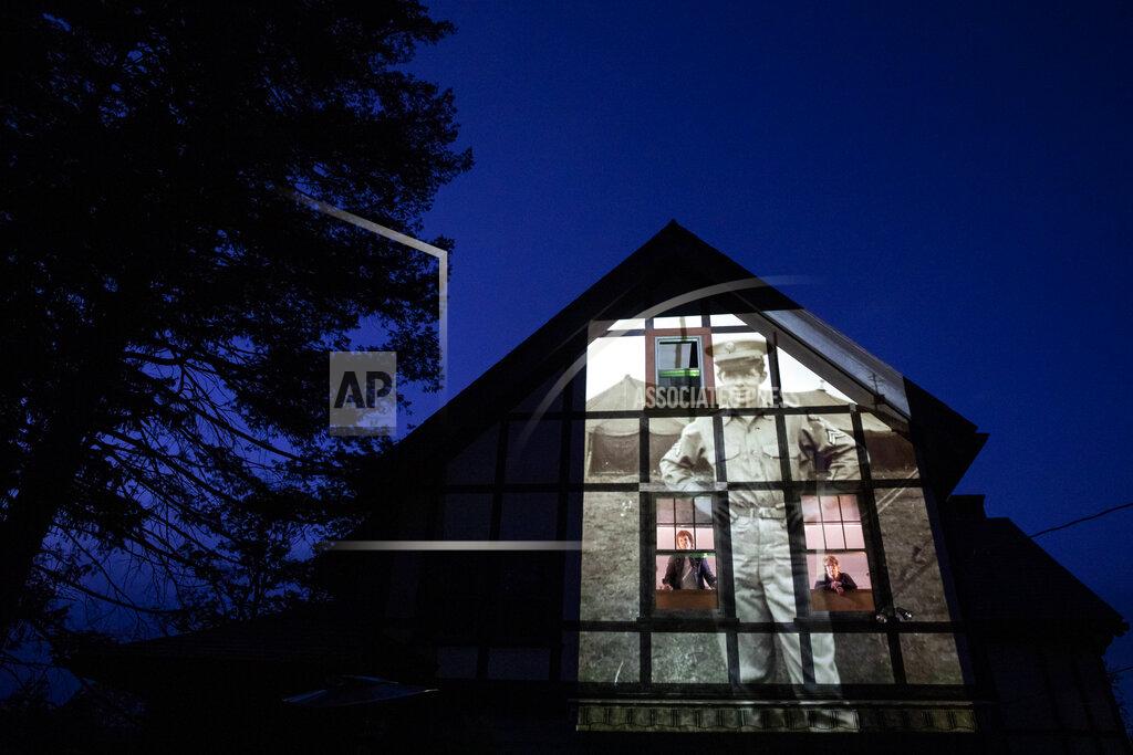 David Goldman An image of veteran Alfred Healy is projected onto the home of his daughter, Eileen Driscoll, left, as she looks out the window with her sister, Patricia Creran, in Holyoke, Mass., Thursday, May 7, 2020. Healy, a U.S. Army Korean War veteran and resident of the Soldier's Home in Holyoke, Mass., died from the COVID-19 virus at the age of 91. Seeking to capture moments of private mourning at a time of global isolation, the photographer used a projector to cast large images of veterans on to the homes as their loved ones are struggling to honor them during a lockdown that has sidelined many funeral traditions. (AP Photo/David Goldman)