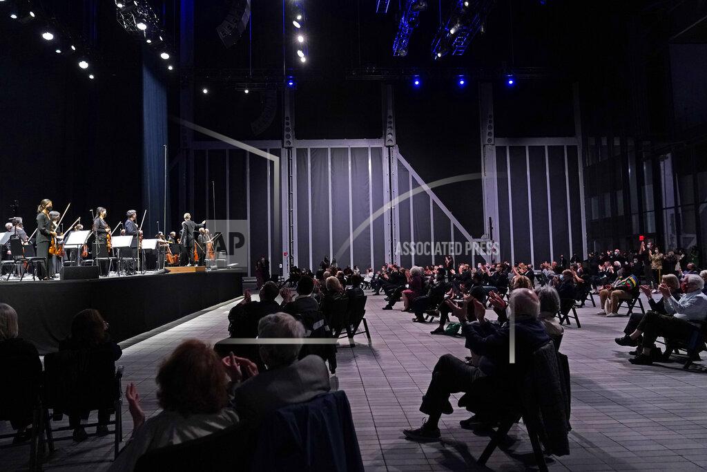 Kathy Willens CORRECTS SPELLING OF FIRST NAME TO ESA INSTEAD OF ESSA - A live audience of spectators applauds as members of the New York Philharmonic stand after the orchestra performed a live concert together for the first time since March 10, 2020, at The Shed in Hudson Yards, Wednesday, April 14, 2021, in New York. Esa-Pekka Salonen, music director of the San Francisco Symphony and principal conductor of London's Philharmonia Orchestra, gestures from the stage. (AP Photo/Kathy Willens)