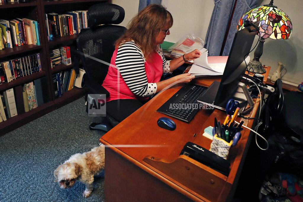 Ellen Boudreau works remotely at her home, Tuesday, Aug. 4, 2020, in Manchester, N.H. Boudreau, who was laid off from her full-time bookkeeping job for six weeks as the COVID-19 virus pandemic shutdown much of the area businesses, now works less than 10 hours per week with that employer, while also taking on a part-time, temporary worker job at another office. Walking out from behind her desk is her 15-year-old dog "Happy Go Lucky". (AP Photo/Charles Krupa)