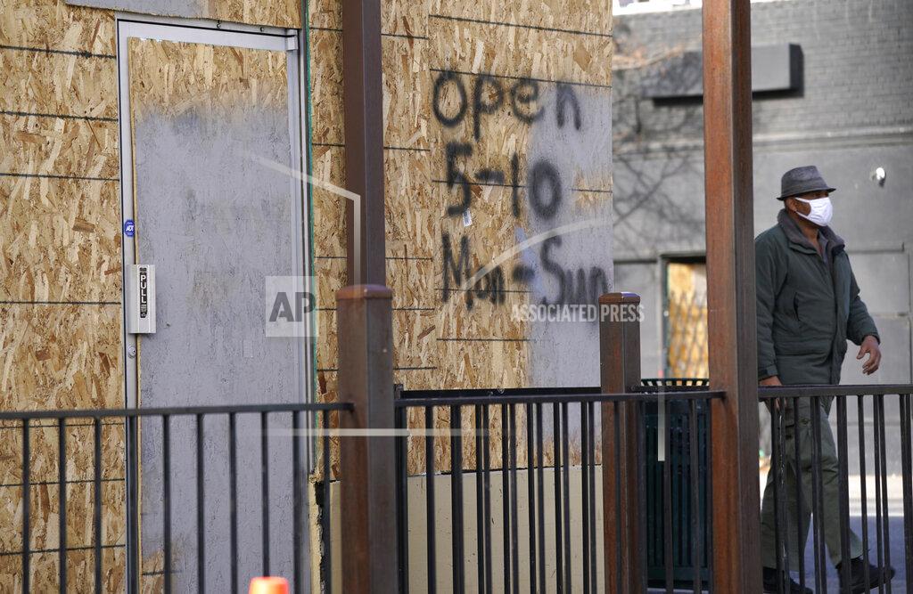 David Zalubowski A pedestrain in a face mask walks past a restaurant with boarded-up windows on which hours of operation are listed along East Colfax Avenue Monday, Dec. 21, 2020, in downtown Denver. (AP Photo/David Zalubowski)