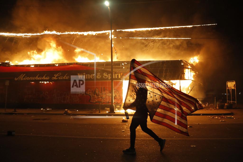 Julio Cortez A protester carries the carries a U.S. flag upside, a sign of distress, next to a burning building Thursday, May 28, 2020, in Minneapolis. Protests over the death of George Floyd, a black man who died in police custody Monday, broke out in Minneapolis for a third straight night. (AP Photo/Julio Cortez)