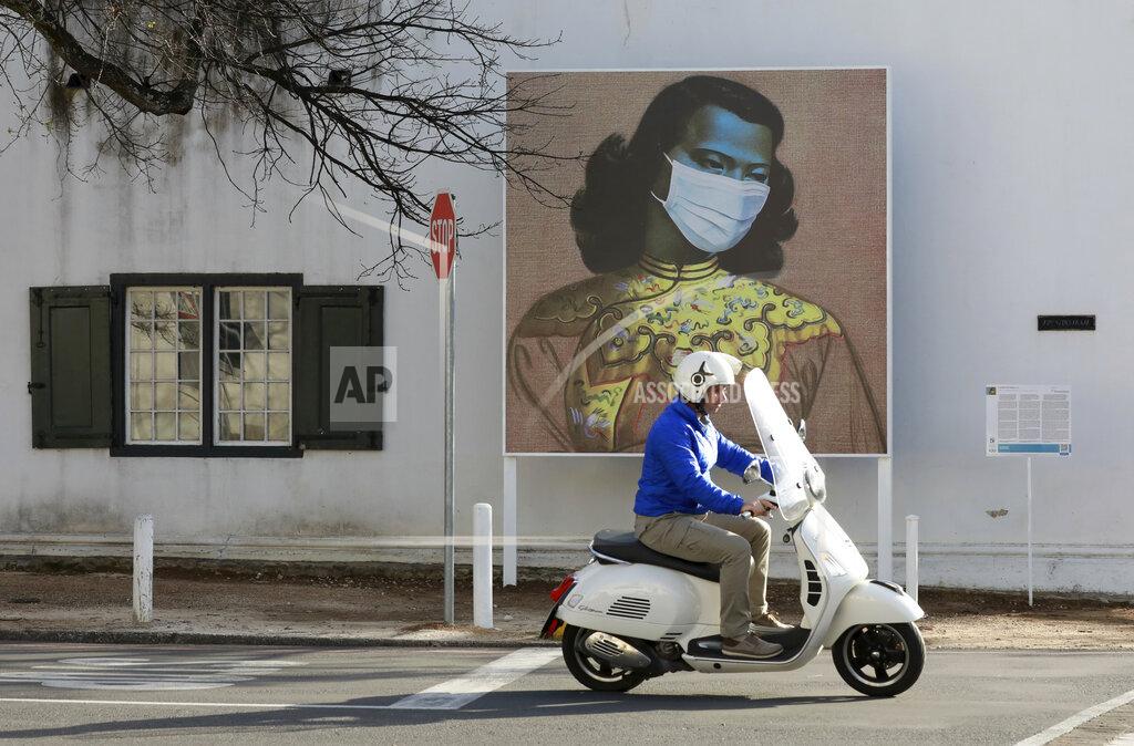 Nardus Engelbrecht A man rides his scooter in front of a marked version of Vladimir Tretchikoff's "The Chinese Girl, The Green Lady" painting in Stellenbosch, South Africa, Thursday Aug. 13, 2020. The public art is part of a series called Masked Masterpieces of masks superimposed on famous South African artworks to support Stellenbosch University students impacted by Covid-19. ( AP Photo/Nardus Engelbrecht)