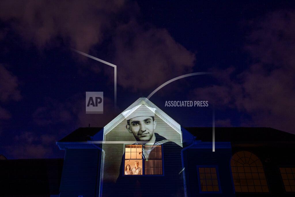 David Goldman An image of veteran Harry Malandrinos is projected onto the home of his son, Paul Malandrinos, as he looks out a window with his wife, Cheryl, in Wilbraham, Mass., Saturday, May 16, 2020. Malandrinos, a U.S. Navy veteran and resident of the Soldier's Home in Holyoke, Mass., died from the COVID-19 virus at the age of 89. Seeking to capture moments of private mourning at a time of global isolation, the photographer used a projector to cast large images of veterans on to the homes as their loved ones are struggling to honor them during a lockdown that has sidelined many funeral traditions. (AP Photo/David Goldman)