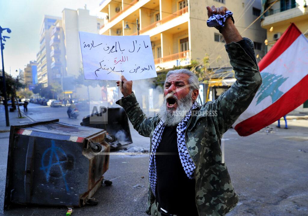 Hussein Malla A protester shouts slogans in front of burning garbage containers used to block a main road during a protest against the increase in prices of consumer goods and the crash of the local currency, in Beirut, Lebanon, Tuesday, March 16, 2021. The Arabic placard reads:"Salameh (the Central Bank governor) is still at the bank while the lira is dead." (AP Photo/Hussein Malla)