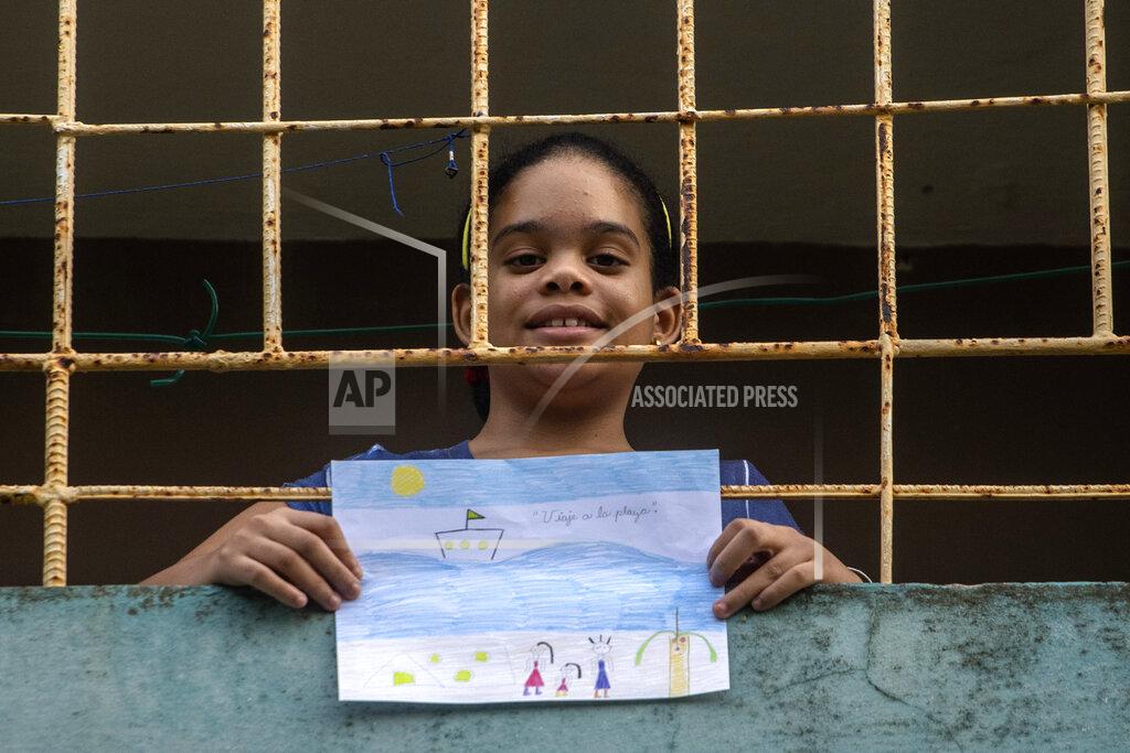 Ramon Espinosa Ana Laura Ramirez Lavandero, 10, holds her drawing in which she expresses her wish to make a trip to the beach, looking out from the balcony of her home in Havana, Cuba, Friday, May 8, 2020. The only time she’s been able to go out in nearly two months has been for an emergency trip to the dentist. Schools are closed, and because many people in Cuba don’t have internet, the education ministry is broadcasting lessons on state television. (AP Photo/Ramon Espinosa )