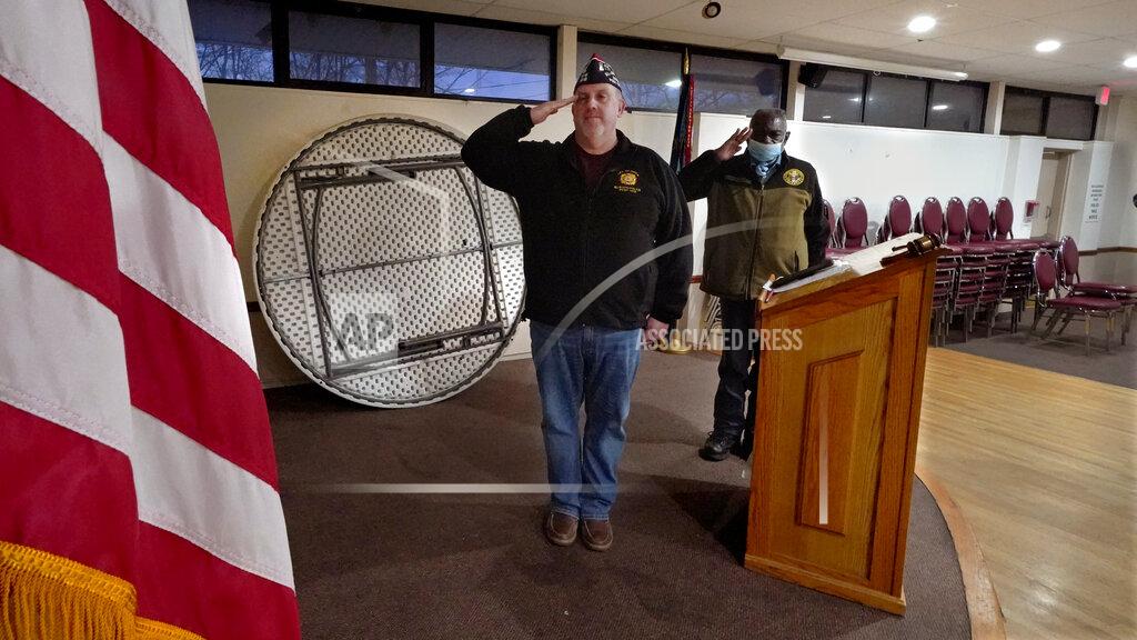 Charles Krupa With tables and chairs stacked against the wall of their rental hall space, Craig DeOld, commander at Veterans of Foreign War Post #1018, center, and Board of Directors member Grady Durden, right, salute the flag at the start of a virtual post meeting, Monday, March 15, 2021, in Boston. Local bars and halls run by VFW and American Legion posts have fallen on hard times during the coronavirus pandemic. Organizers say many risk permanent closure after states ordered them, like other bars and halls, to shutter last spring. (AP Photo/Charles Krupa)