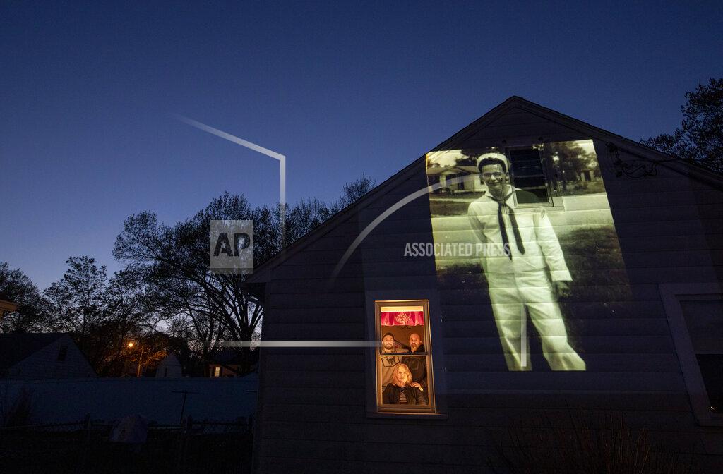 David Goldman An image of veteran James Mandeville is projected onto the home of his daughter, Laurie Mandeville Beaudette, as she looks out a window with her son, Kyle, left, and husband, Mike, in Springfield, Mass., Tuesday, May 12, 2020. Mandeville, a U.S. Navy veteran and resident of the Soldier's Home in Holyoke, Mass., died from the COVID-19 virus at the age of 83. Seeking to capture moments of private mourning at a time of global isolation, the photographer used a projector to cast large images of veterans on to the homes as their loved ones are struggling to honor them during a lockdown that has sidelined many funeral traditions. (AP Photo/David Goldman)
