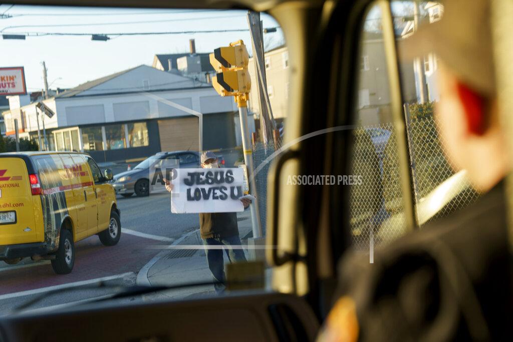 A man holds a sign which reads, "Jesus Loves U" at an intersection as Ryan Bradley, right, a firefighter and the city's emergency medical services coordinator, rides by in an ambulance in Central Falls, R.I., Monday, March 29, 2021. Bradley's family, French-Canadian immigrants on one side and Irish on the other, goes back in Central Falls for generations. He's proud of the city's long immigrant tradition. "It's one thing I loved about Central Falls growing up here: It's the best best place to learn about the world without having to travel very far." (AP Photo/David Goldman)