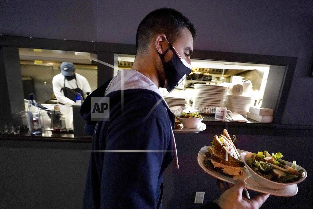 Steven Senne Bartender Denis Angelov, of Provincetown, Mass., left, carries plates of food from the kitchen, behind, at Tin Pan Alley restaurant, Tuesday, April 6, 2021, in Provincetown. Hotels, restaurants and other businesses in tourist destinations are warning that hiring challenges during the coronavirus pandemic could force them to pare back operating hours or curtail services just as they’re eyeing a bounce-back summer. (AP Photo/Steven Senne)