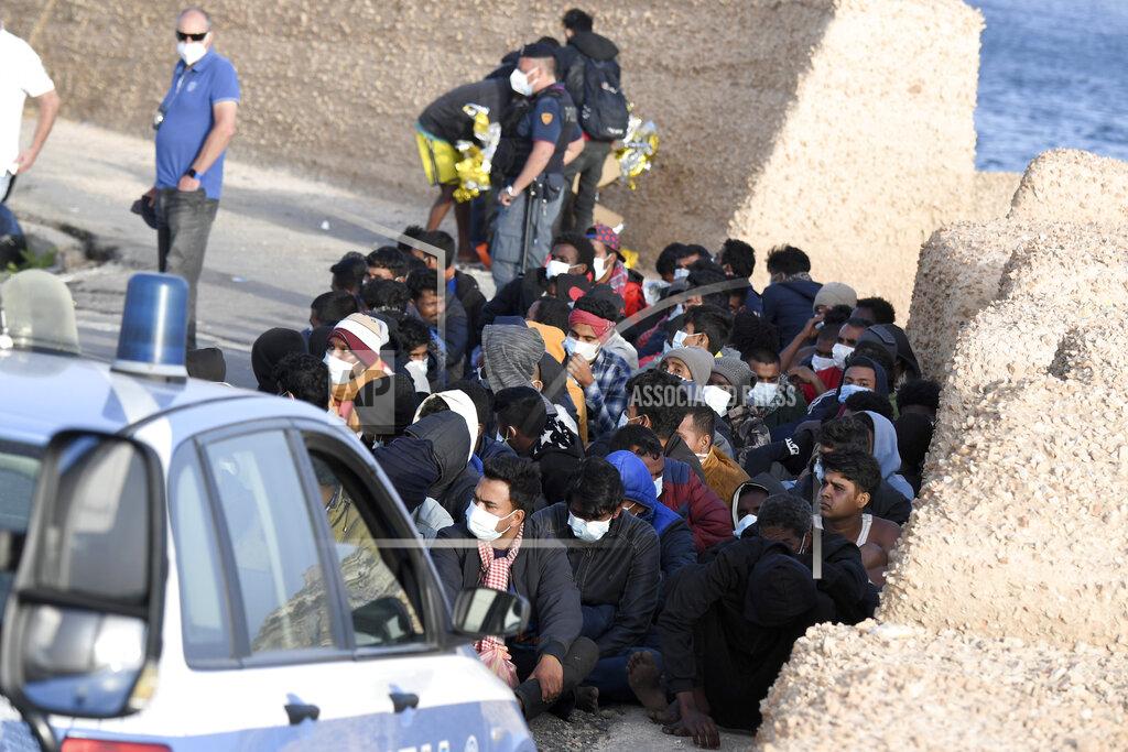 Salvatore Cavalli Migrants wearing face masks to curb the spread of COVID-19 sit at a pier as Italian police officers stand by, on the Sicilian island of Lampedusa, southern Italy, Monday, May 10, 2021. Several hundred more migrants reached the tiny Italian island before dawn on Monday, swelling to past 2,100 the number of arrivals in around 24 hours and fueling calls from across the political spectrum for the Italian government to strengthen its migration policies. (AP Photo/Salvatore Cavalli)