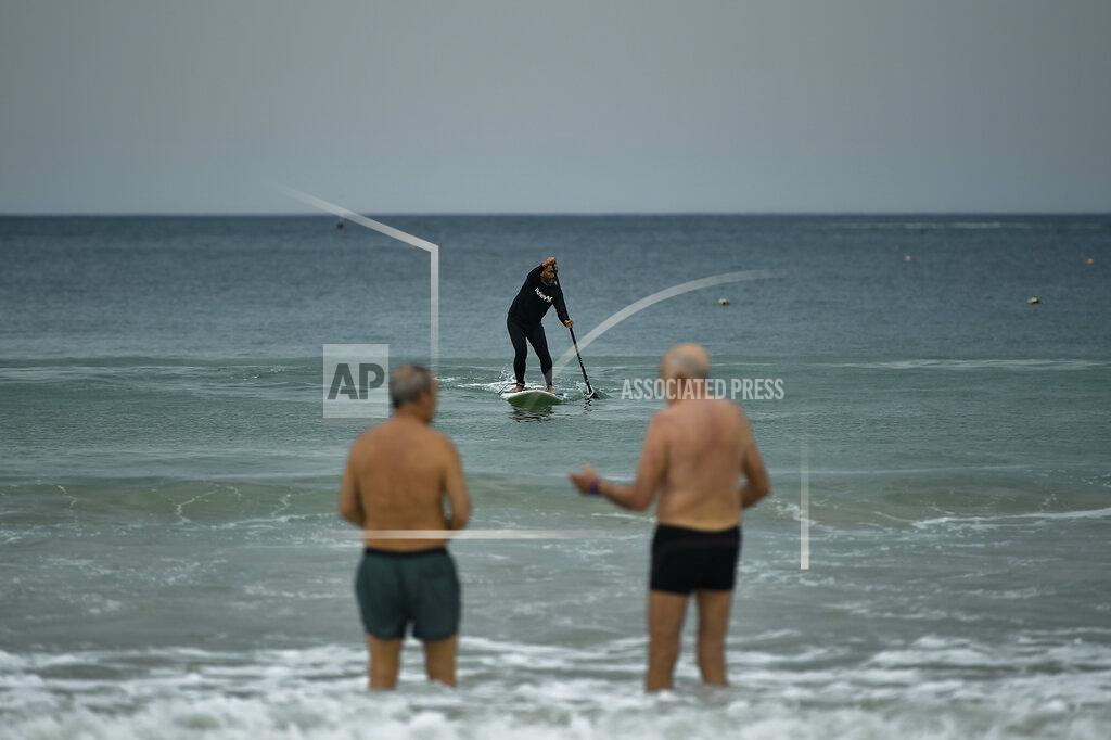Alvaro Barrientos A surferin the sea, while two old men talks, at La Concha beach, after lockdown restrictions were lifted, in San Sebastian, northern Spain, Sunday, May 9, 2021. Impromptu street celebrations erupted across Spain as the clock struck midnight on Saturday, when a six-month-long national state of emergency to contain the spread of coronavirus ended and many nighttime curfews were lifted. (AP Photo/Alvaro Barrientos)