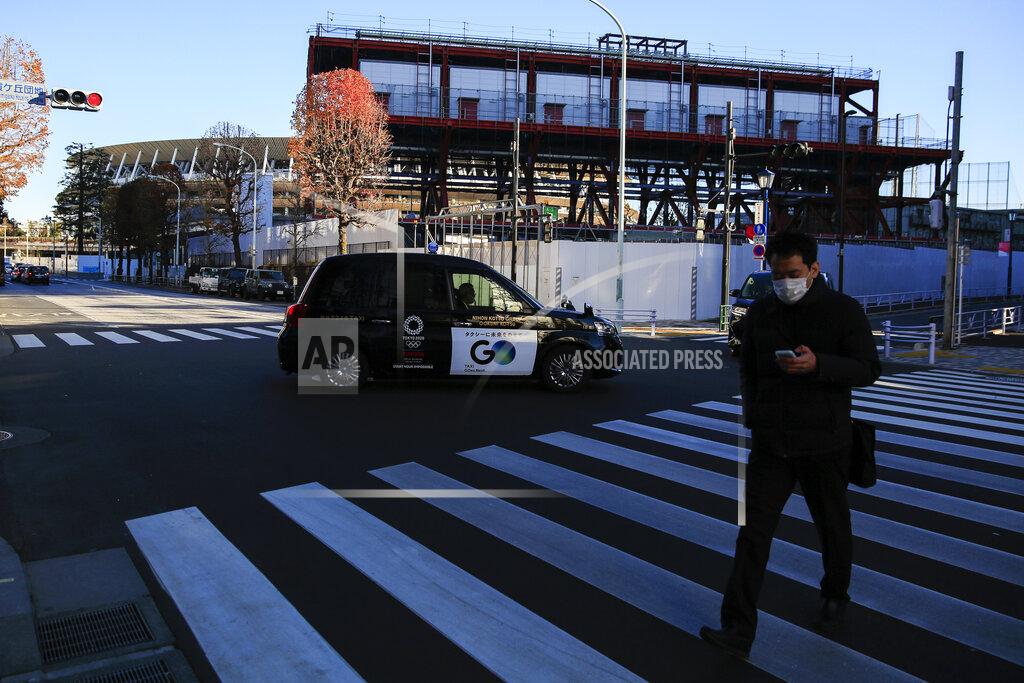 Kiichiro Sato A man walks across a street as a massive steel structure of the TV Tower, that houses studios for live broadcasts of Tokyo 2020, in front of National Stadium, where opening ceremony and many other events are planned for postponed Tokyo 2020 Olympics, is seen from a rooftop observation deck Thursday, Jan. 21, 2021, in Tokyo. The postponed Tokyo Olympics are to open in just six months. Local organizers and the International Olympic Committee say they will go ahead on July 23. But it’s still unclear how this will happen with virus cases surging in Tokyo and elsewhere around the globe. (AP Photo/Kiichiro Sato)