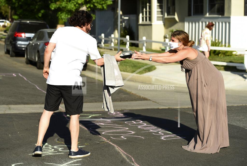 Chris Pizzello In this May 9, 2020 photo, musician Adam Chester, left, receives carrot cake cupcakes from neighbor Caryn Adams over a social-distancing chalk line before Chester's weekly neighborhood concert in the Sherman Oaks section of Los Angeles. Normally, Chester is a surrogate Elton John, who sings and plays the rock superstar's parts at rehearsals. With that work on hold, Chester has been giving concerts to his neighbors from a safe social distance in front of his house. (AP Photo/Chris Pizzello)