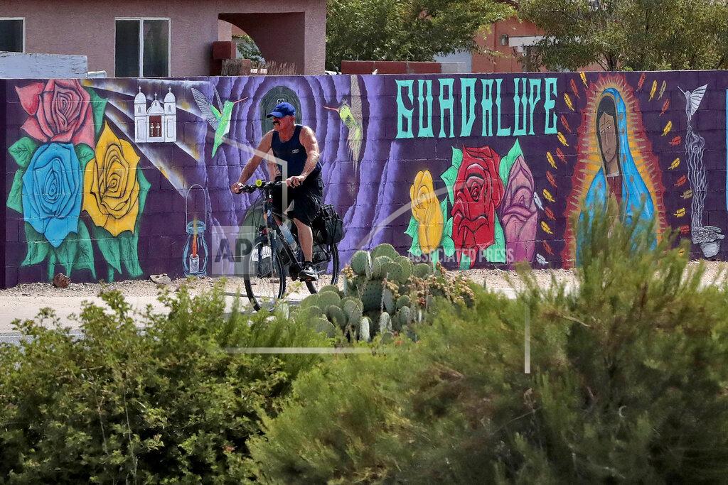 In this Saturday, June 13, 2020, photo, a man cycles past a mural in Guadalupe, Ariz. As the coronavirus spreads deeper across America, it's ravaging through the homes and communities of Latinos from the suburbs of the nation's capital to the farm fields of Florida to the sprawling suburbs of Phoenix and countless communities in between. (AP Photo/Matt York)