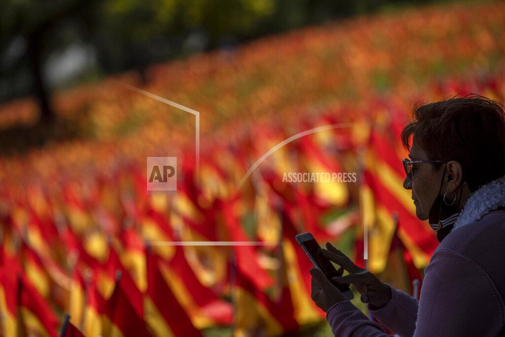 Manu Fernandez A woman sits near to the Spanish flags placed in memory of coronavirus (COVID-19) victims, in Madrid, Spain, Sunday, Sept. 27, 2020. An association of families of coronavirus victims has planted what it says are 53,000 small Spanish flags in a Madrid park to honor the dead of the pandemic. (AP Photo/Manu Fernandez)