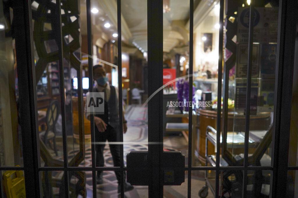 Andrew Medichini A staffer stands behind the bars of a closed restaurant, in Rome, Monday, Oct. 26, 2020. For at least the next month, people outdoors except for small children must now wear masks in all of Italy, gyms, cinemas and movie theaters will be closed, ski slopes are off-limits to all but competitive skiers and cafes and restaurants must shut down in early evenings, under a decree signed on Sunday by Italian Premier Giuseppe Conte, who ruled against another severe lockdown despite a current surge in COVID-19 infections. (AP Photo/Andrew Medichini)