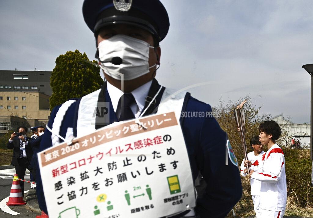 Philip Fong A guard with a sign for coronavirus precautions stands as Japanese Azusa Iwashimizu, right, a member of Japan women's national football team, carries an Olympic Torch during the torch relay grand start outside J-Village National Training Center in Naraha, Fukushima prefecture, northeastern Japan, Thursday, March 25, 2021. The torch relay for the postponed Tokyo Olympics began its 121-day journey across Japan on Thursday and is headed toward the opening ceremony in Tokyo on July 23. (Philip Fong/Pool Photo via AP)