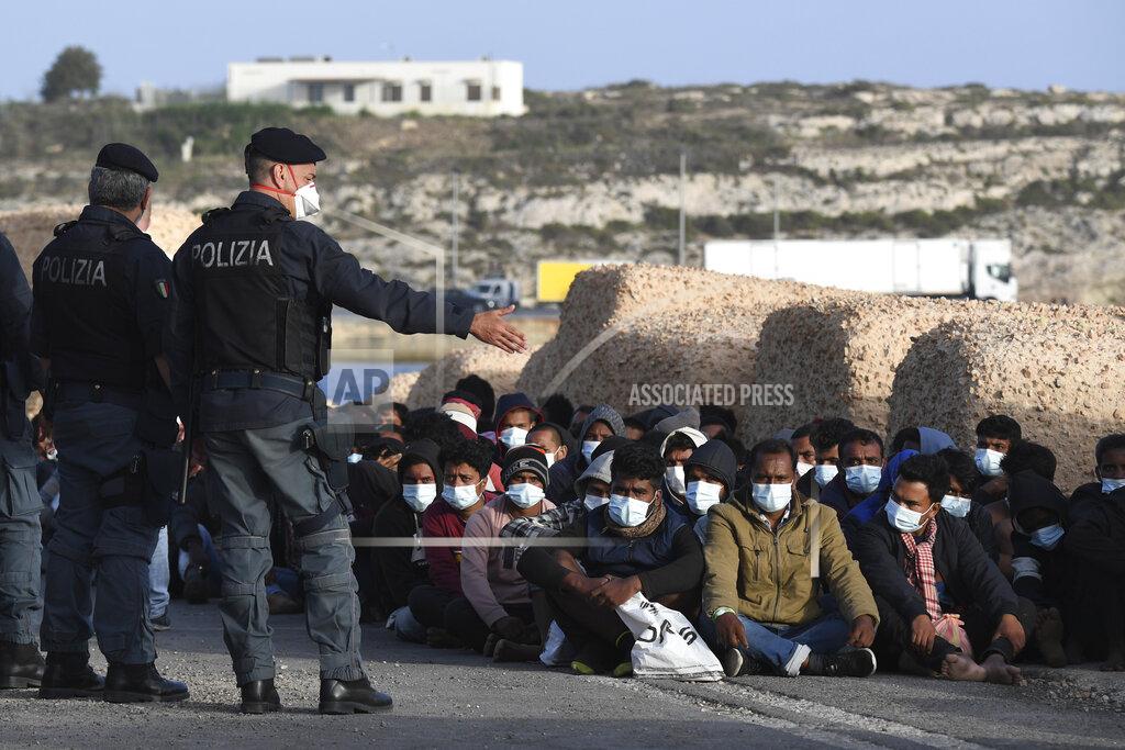 Salvatore Cavalli Migrants wearing face masks to curb the spread of COVID-19 sit at a pier as Italian police officers stand by, on the Sicilian island of Lampedusa, southern Italy, Monday, May 10, 2021. Several hundred more migrants reached the tiny Italian island before dawn on Monday, swelling to past 2,100 the number of arrivals in around 24 hours and fueling calls from across the political spectrum for the Italian government to strengthen its migration policies. (AP Photo/Salvatore Cavalli)