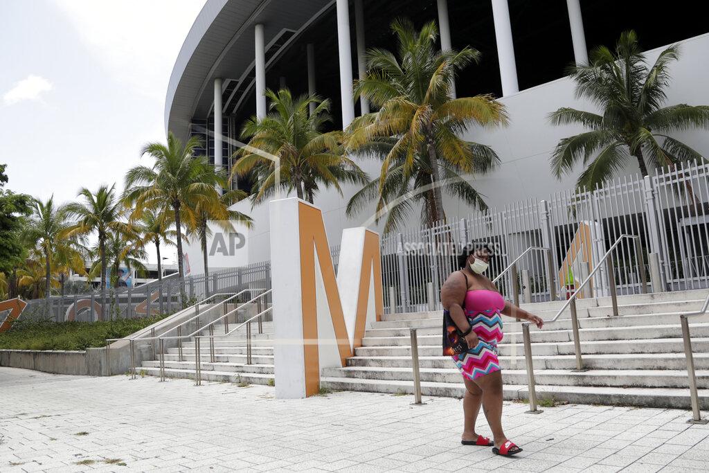 Lynne Sladky A woman wearing a protective face covering walks past Marlins Park, Monday, July 27, 2020, in Miami. The Marlins home opener against the Baltimore Orioles on Monday night has been postponed as the Marlins deal with a coronavirus outbreak that stranded them in Philadelphia. (AP Photo/Lynne Sladky)