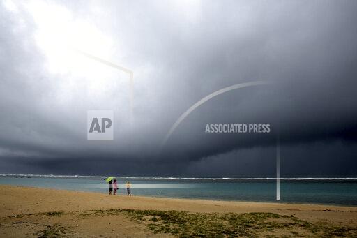 People hold umbrellas as it begins to rain on an otherwise empty beach in Honolulu on Monday, Dec. 6, 2021. A strong storm packing high winds and extremely heavy rain flooded roads and downed power lines and tree branches across Hawaii, with officials warning Monday of potentially worse conditions ahead. (AP Photo/Caleb Jones)