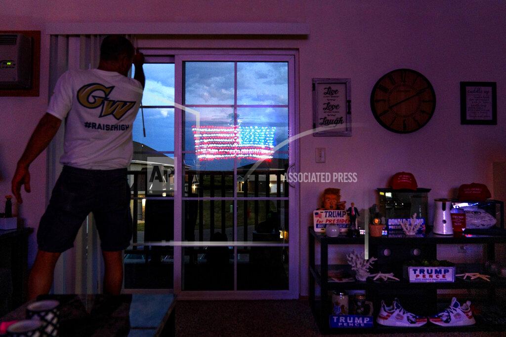 David Goldman An illuminated American flag is reflected in the balcony door of Scott Rice's apartment in Appleton, Wis., Aug. 20, 2020. Rice reveres the president the way Wisconsin loves the Green Bay Packers. He has painted "T-R-U-M-P" on his lawn, spelled it out with Christmas lights on his roof and painted it on his steel-toed shoes. (AP Photo/David Goldman)