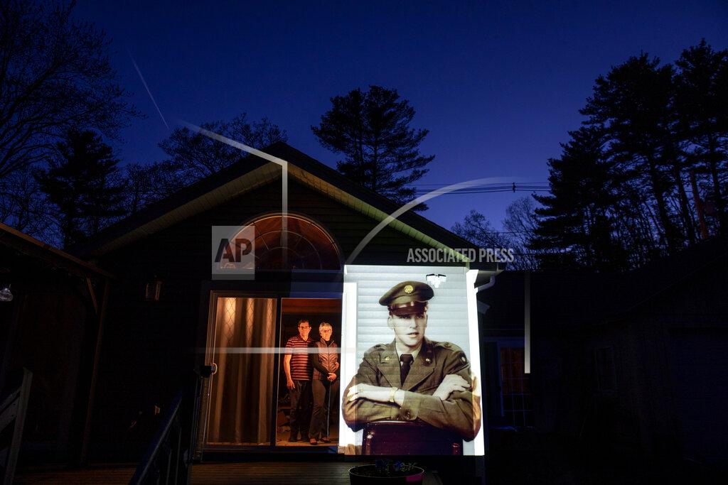 David Goldman An image of veteran Roy Benson is projected onto the home of his daughter, Robin Benson Wilson, left, as she looks out a doorway with her husband, Donald, in Holland, Mass., Wednesday, May 13, 2020. Benson, a U.S. Army veteran and resident of the Soldier's Home in Holyoke, Mass., died from the COVID-19 virus at the age of 88. Seeking to capture moments of private mourning at a time of global isolation, the photographer used a projector to cast large images of veterans on to the homes as their loved ones are struggling to honor them during a lockdown that has sidelined many funeral traditions. (AP Photo/David Goldman)
