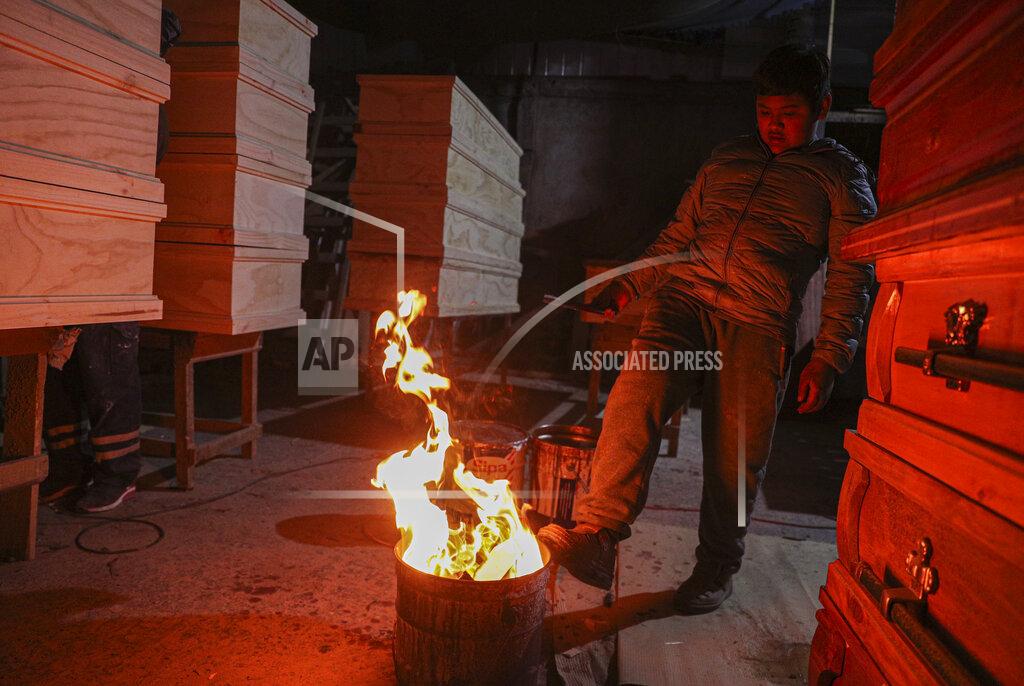 Esteban Felix Cesar Ambrosio warms his feet on a fire while his father works in the finishing details of a production line of coffins at the Bergut Funeral Services' factory in Santiago, Chile, Thursday, June 18, 2020. The coffin production has had to increase up to 120%, according to Nicolas Bergerie, owner of the factory. His more basic coffin model is called the COVID model and is made to cope with the increase of deaths during the coronavirus pandemic. (AP Photo/Esteban Felix)