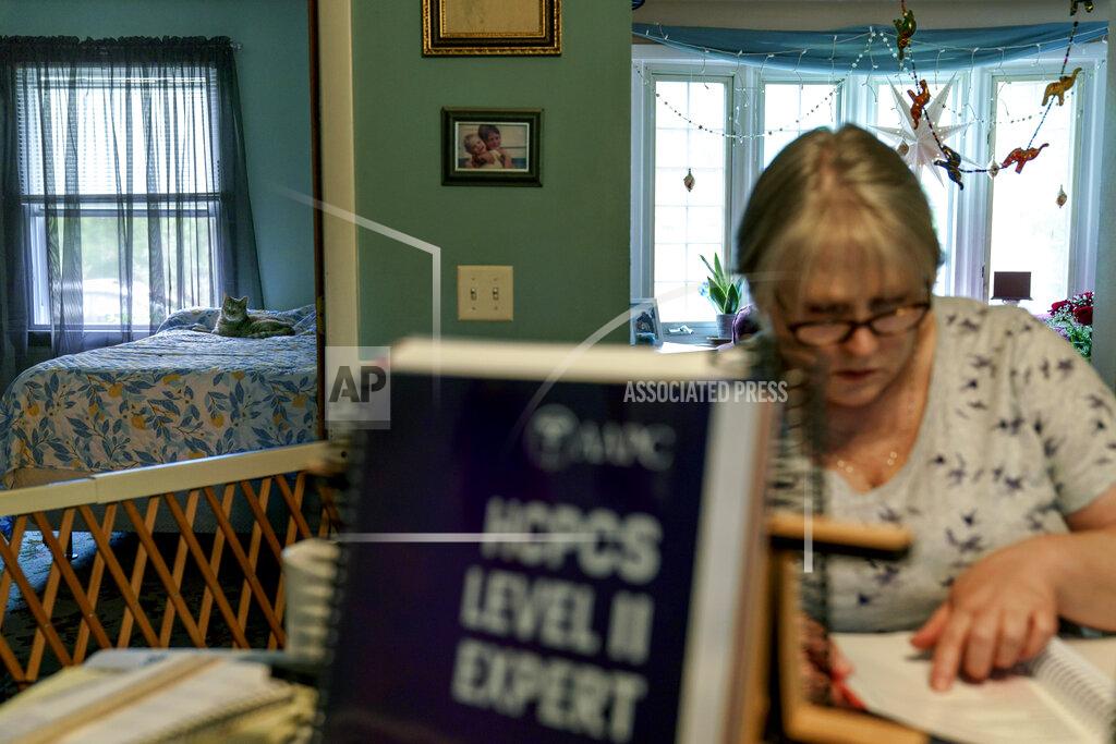 David Goldman Ellen Booth, 57, studies at her kitchen table to become a certified medical coder as her cat, Juji, sits on a bed behind her in Coventry, R.I., Monday, May 17, 2021. When the restaurant she worked for closed last year, Booth said it gave her "the kick I needed." She started a year-long class to learn to be a medical coder. When her unemployment benefits ran out two months ago, she started drawing on her retirement funds. Booth hopes to pass her upcoming exam and soon hit the job market. (AP Photo/David Goldman)