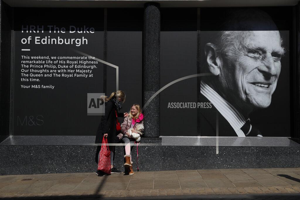 Isabella Disley greets her mother Tara Lynn and her dog Coco next to a portrait of Prince Philip, which adorns a window of a department store in Windsor, England, Friday, April 16, 2021. Prince Philip husband of Britain's Queen Elizabeth II died April 9, aged 99, his funeral will take place Saturday at Windsor Castle in St George's Chapel. (AP Photo/Alastair Grant)