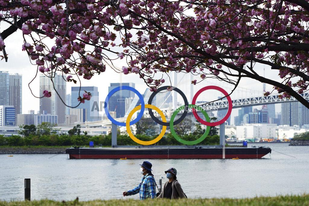 Eugene Hoshiko Under blooming cherry blossoms, people wearing protective masks to help curb the spread of the coronavirus walk with a backdrop of the Olympic rings floating in the water in the Odaiba section Thursday, April 8, 2021, in Tokyo. Many preparations are still up in the air as organizers try to figure out how to hold the postponed games in the middle of a pandemic. (AP Photo/Eugene Hoshiko)